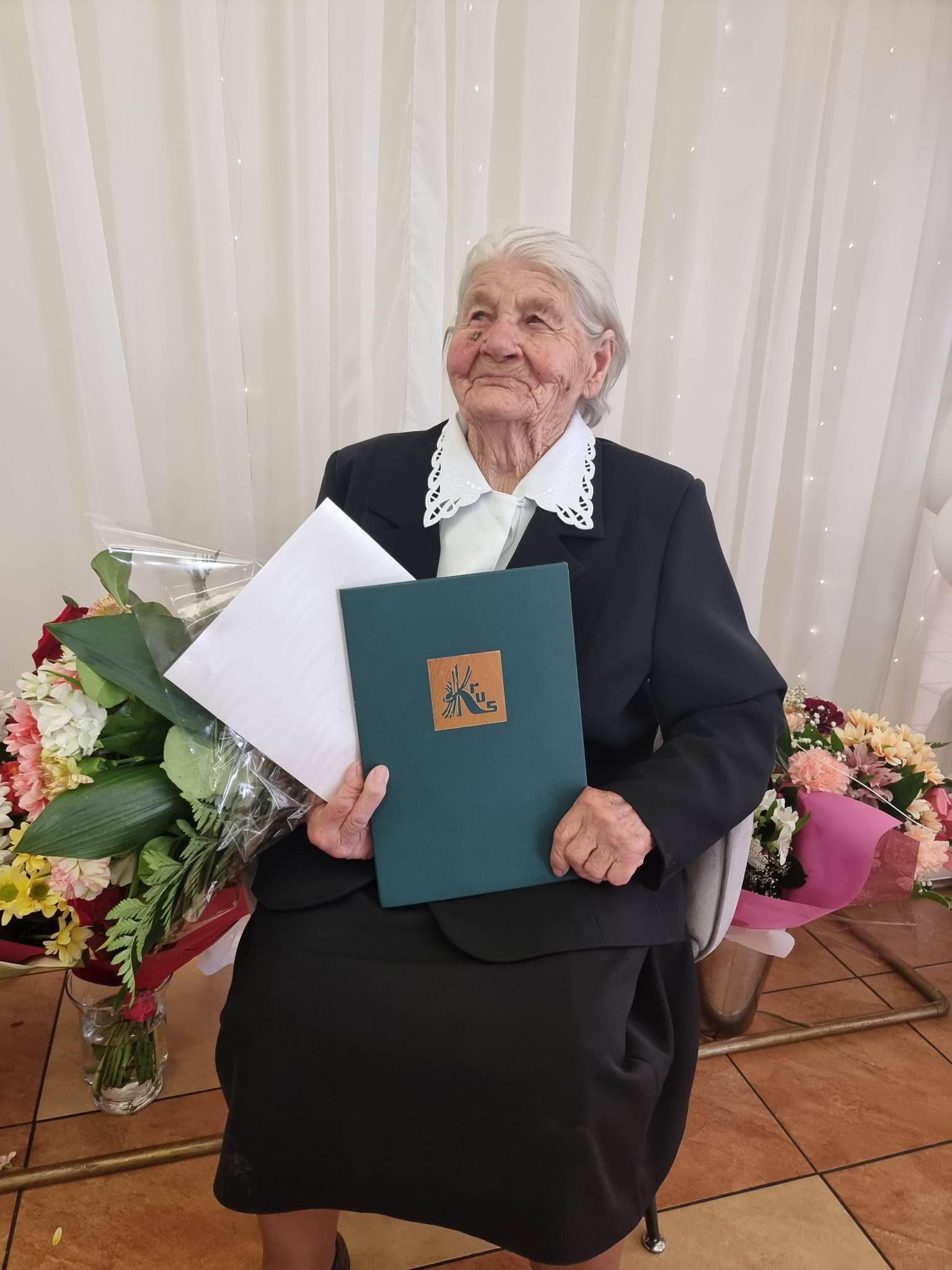 Zdjęcie przedstawia siedzącą jubilakę trzymającą w dłoniach listy gratulacyjne oraz kwiaty